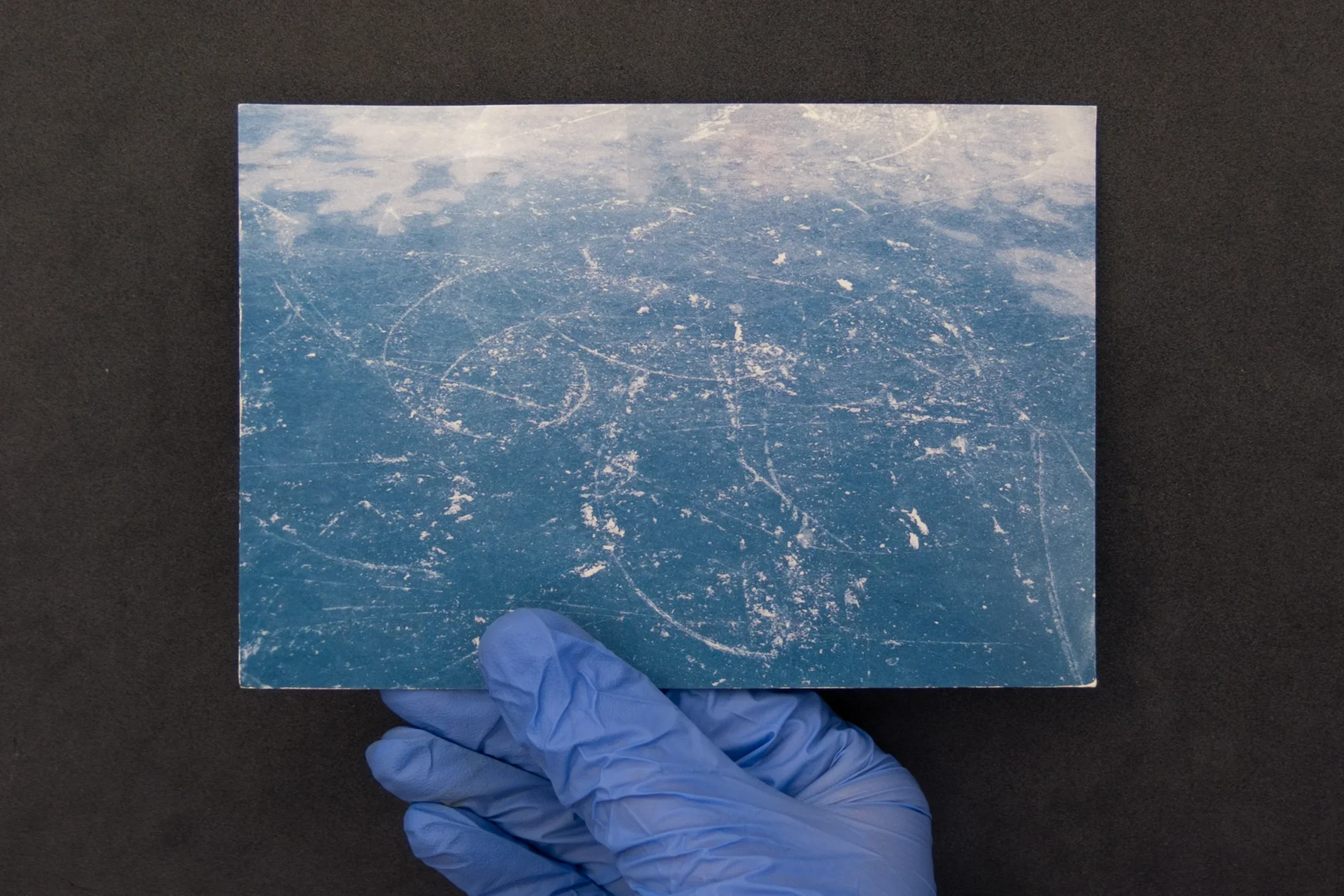 Against a black background, a hand in blue latex-glove holds a postcard showing white curvy lines drawn by skates on blue ice.