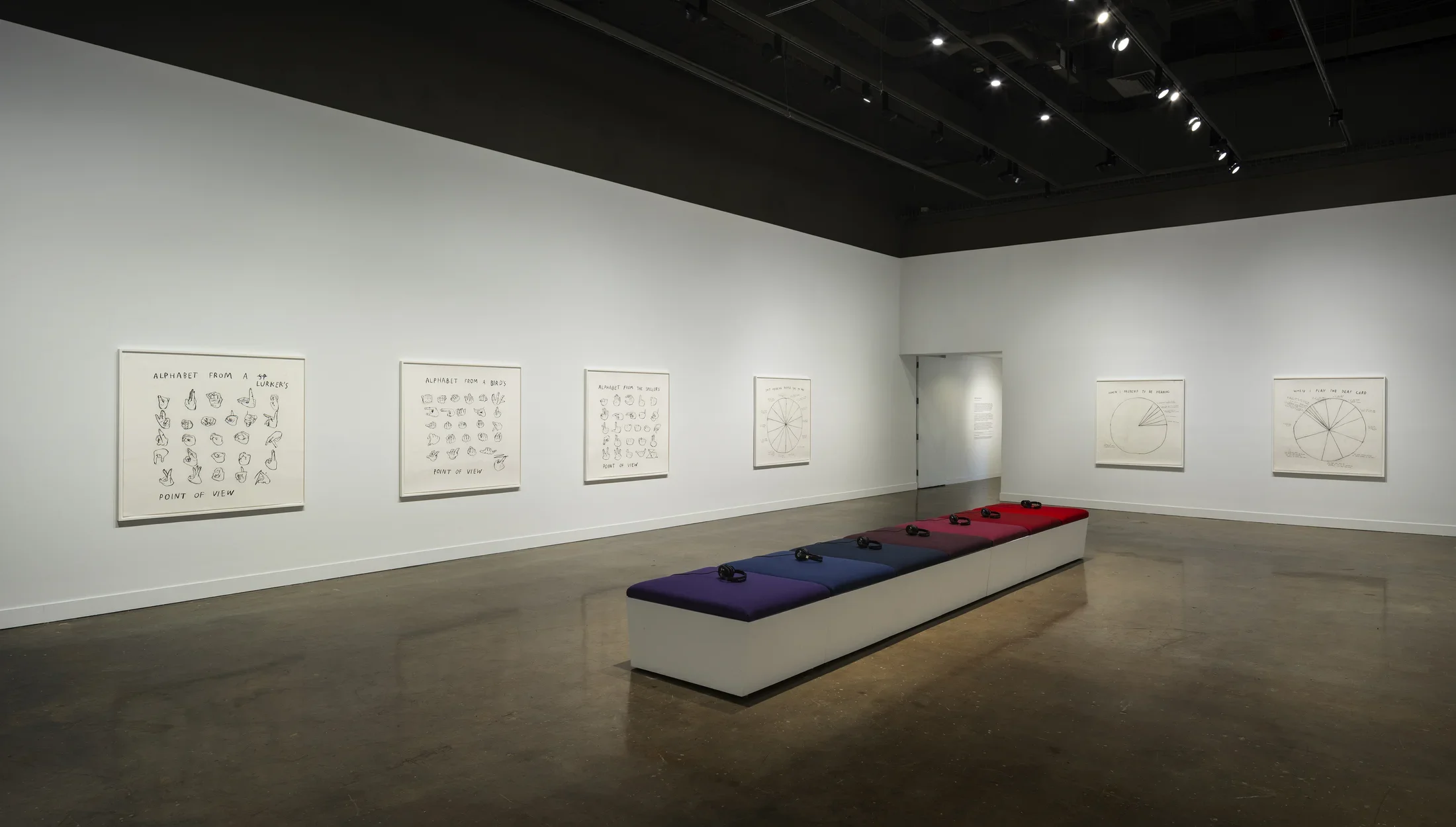 7 headphones on 7 bench cushions ranging from purple to blues to reds sit centered in the gallery with 6 drawings on 2 walls.