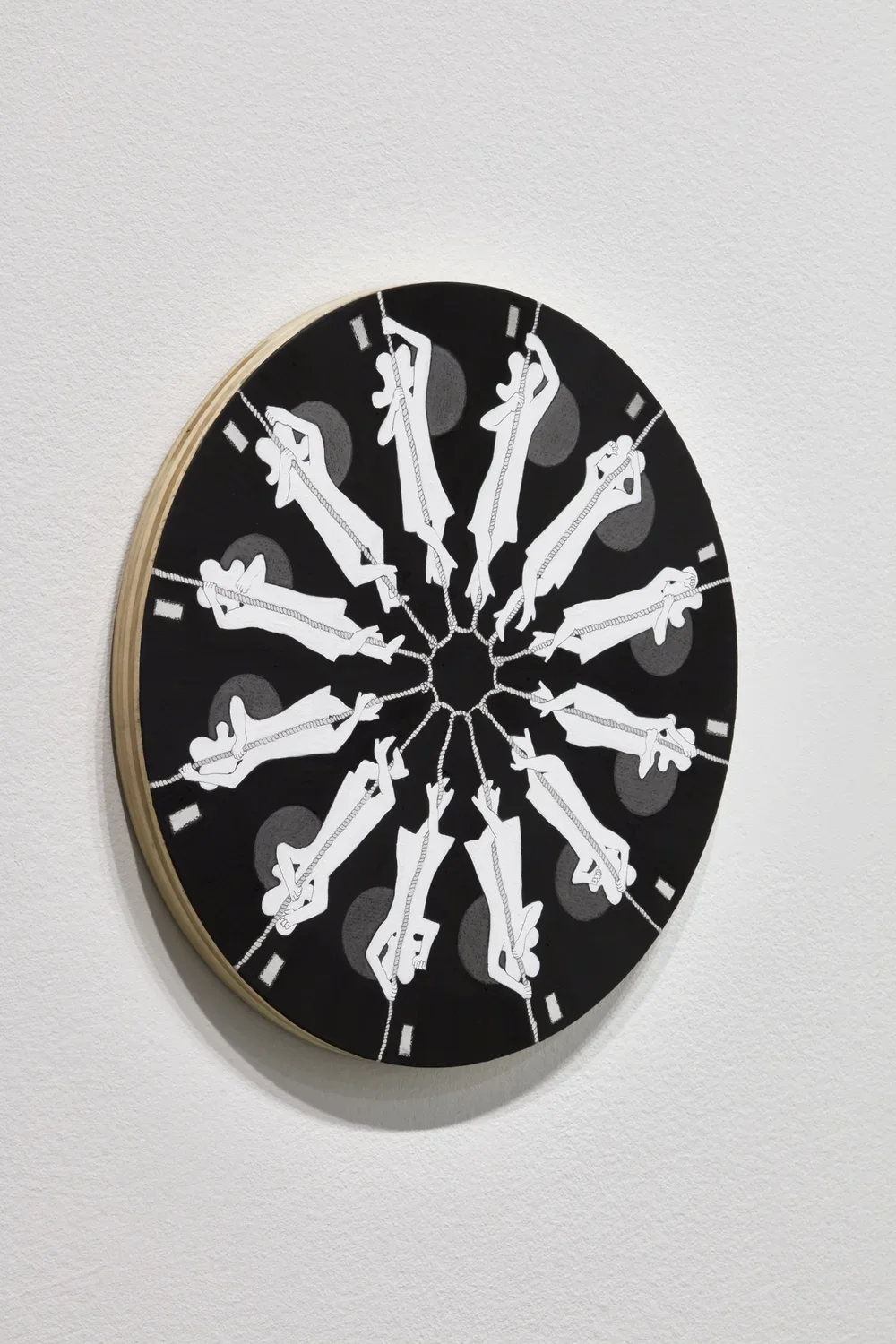 Detail side angle view of small wooden circle affixed to gallery wall. On it, a repeating pattern of white figures climbing a rope against a black background.