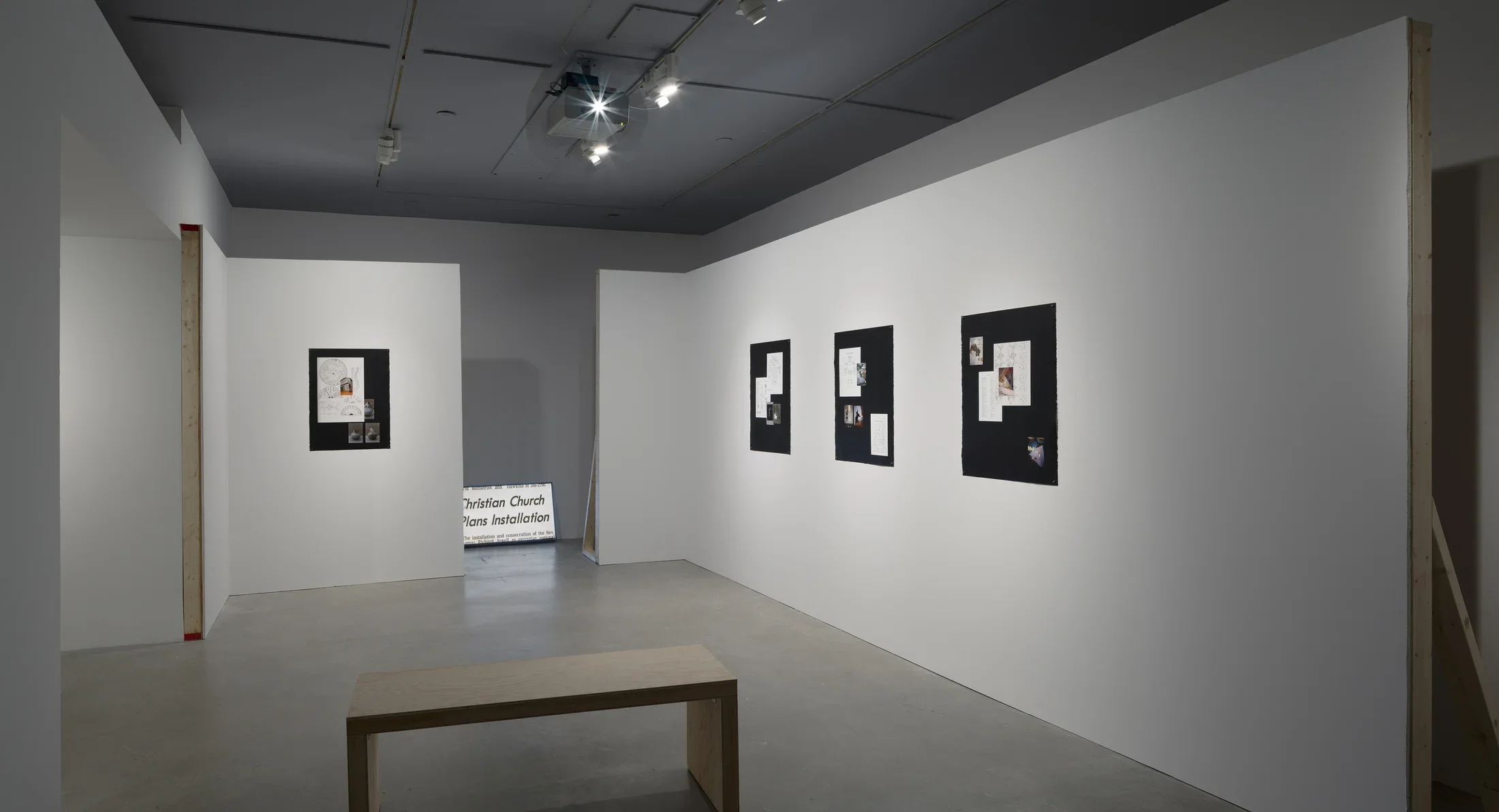 Wood bench in front of 4 paper works mounted on 2 constructed walls and a monitor on the floor between the 2 back walls