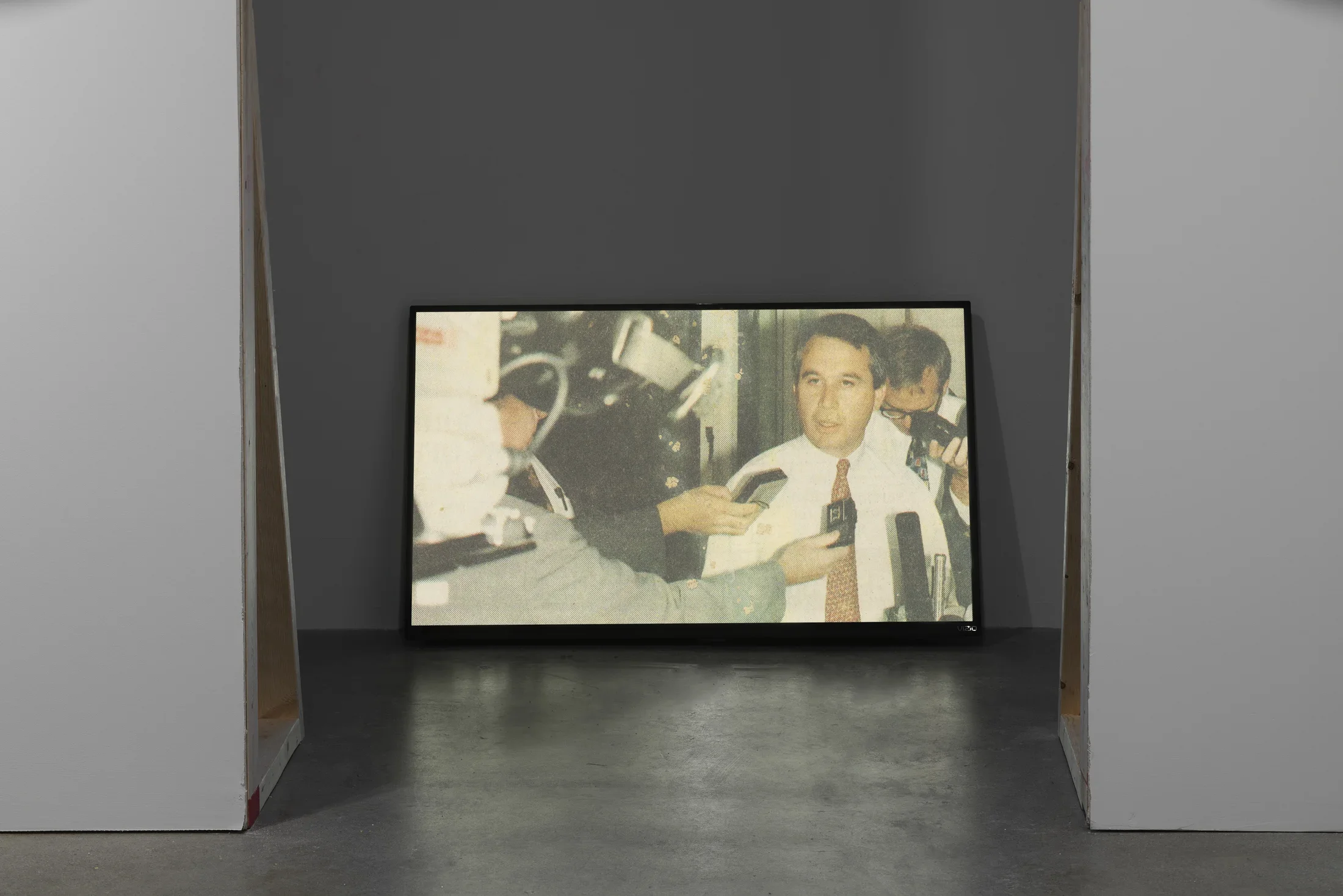 Seen from waist up, a man in shirt and tie speaks into tape recorders and mics in this still shown on a floor monitor.