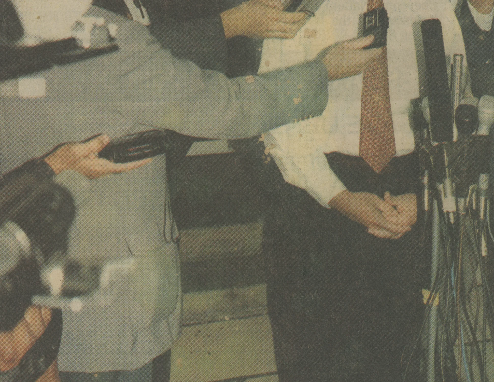 Seen from chest to knee, man in shirt and tie stands behind a cluster of microphones as other men point recorders toward him.