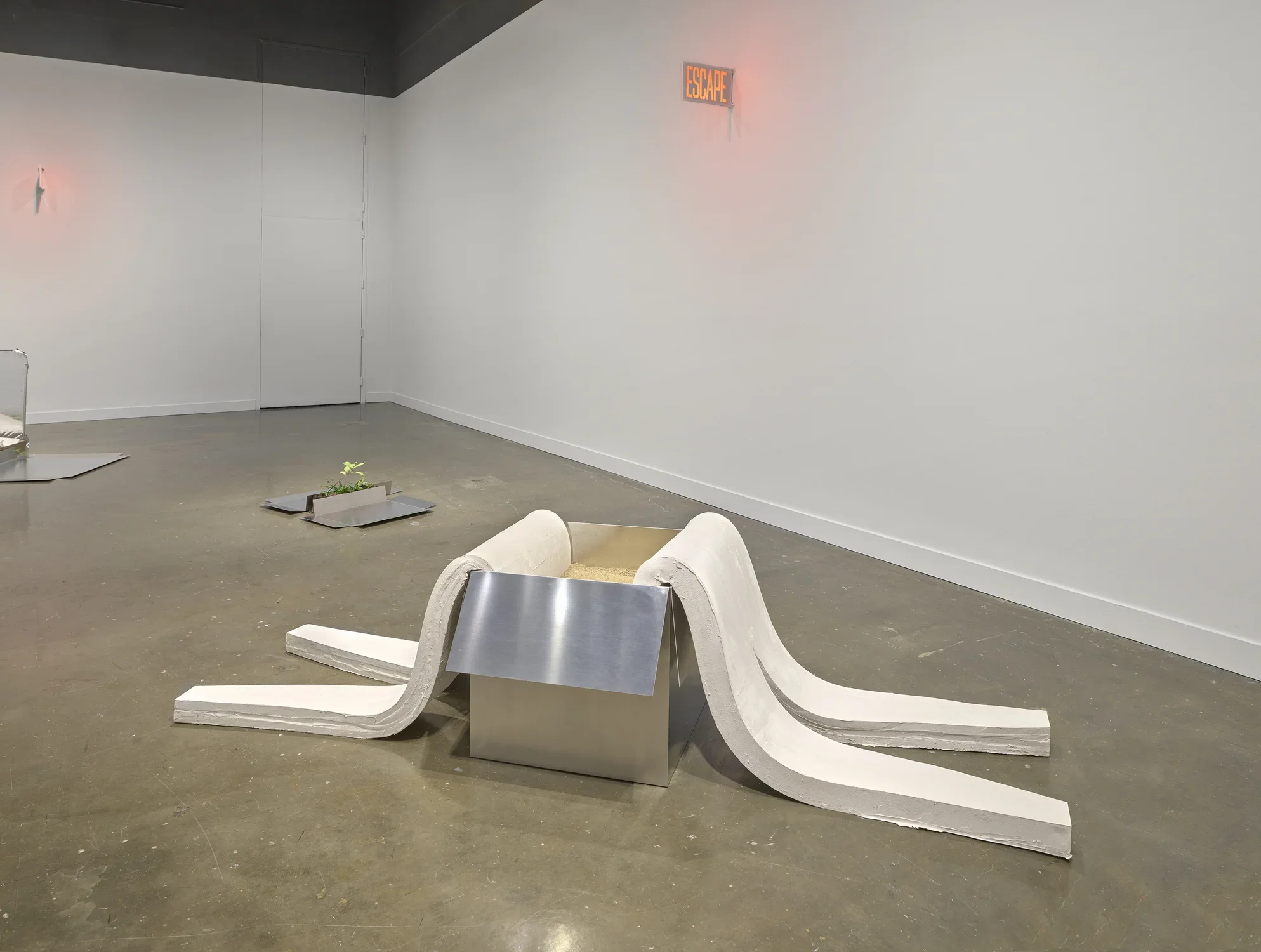 A gallery at the MIT List Visual Arts Center features sculptures consisting of white plaster slumps, aluminum sheets, weeds, and red glowing signs on the walls.