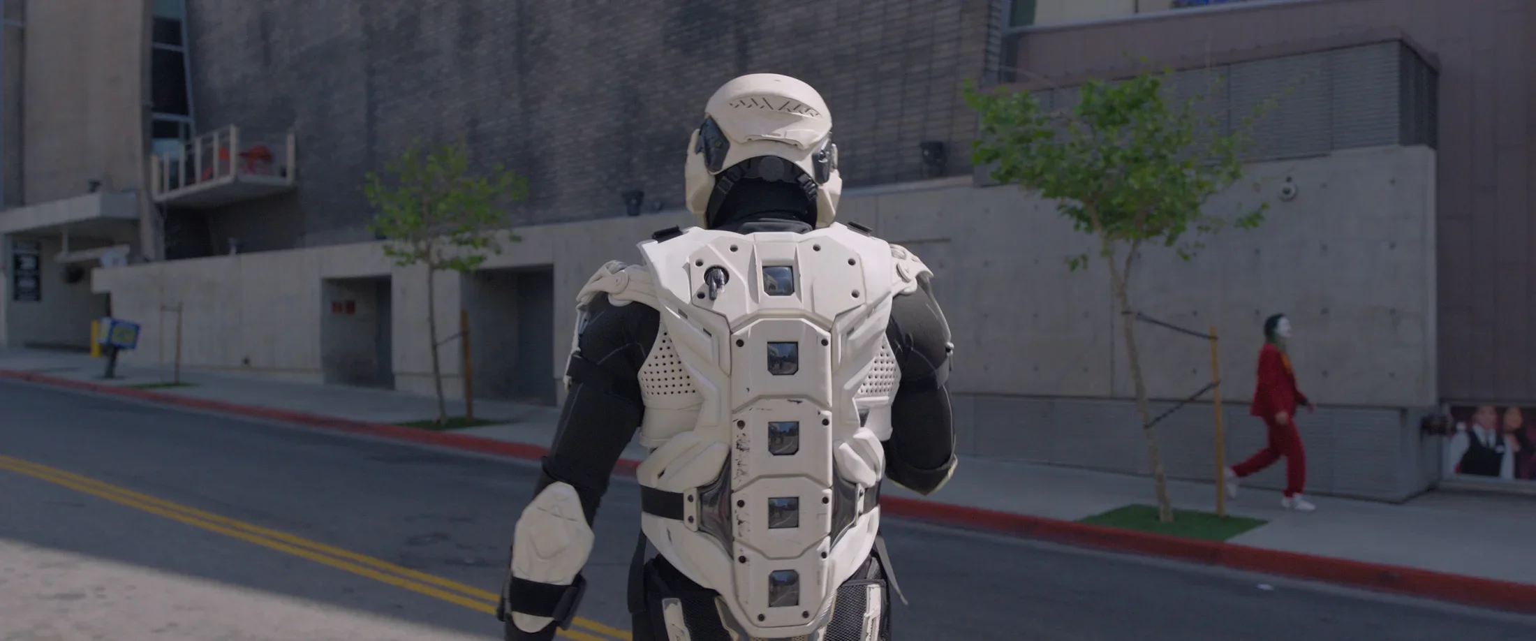 A figure in a black and white storm-trooper-like costume walks down the street with its back turned to the camera. A person in a red suit  green wig and Joker makeup walks on the distant opposing street.
