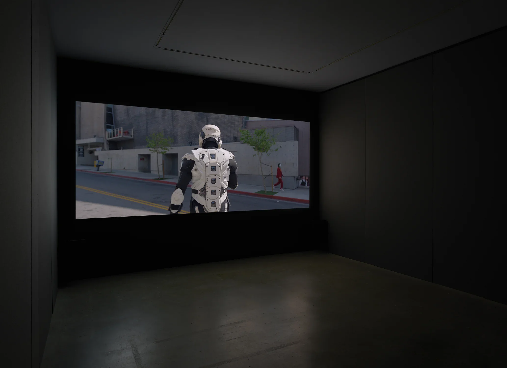 A dimly lit gallery space includes a screening of a storm tropper with its back turned to the screen and a soft bench. 