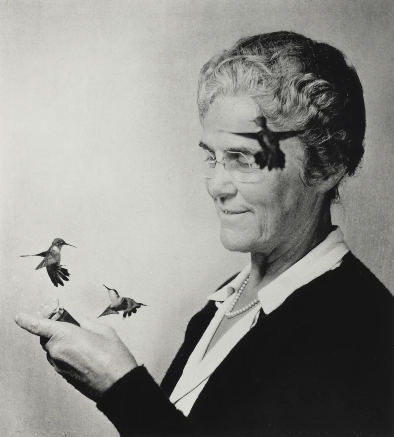 Black and white photograph of an older woman feeding two hummingbirds out of her hands while a third flys near her forehead.