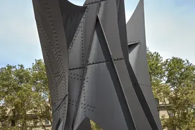 Installation view of large scale steel outdoor sculpture with a blue sky and buildings in the background.