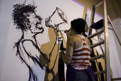 A woman stands on a ladder and paints onto a figure onto a wall in black paint.