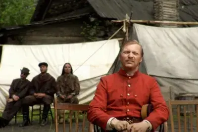 Man in a red shirt sits on a chair in the forefront with 3 people sitting behind him and white tents in the background.