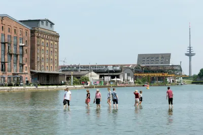 9 people walking in water up to their knees on a shore near buildings.