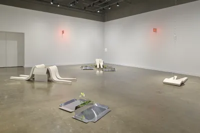 A gallery at the MIT List Visual Arts Center features sculptures consisting of white plaster slumps, aluminum sheets, weeds, and red glowing signs on the walls.