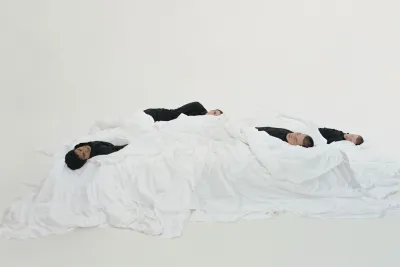 Still of 3 people lying on a mattress wearing black clothing coverd by white sheets