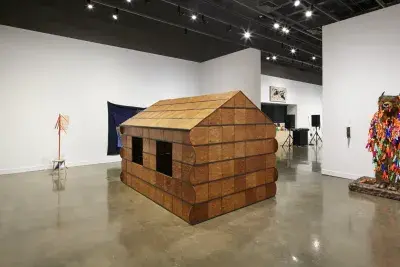 Installation view featuring a wooden cabin sculpture in the foreground