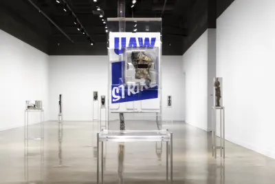 Installation view featuring a work glove in front of a blue and white poster that reads “UAW” on strike encased in a block of resin on a metal stand.