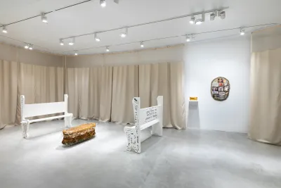 Two white church pews with text on sides and backs frame a sculpture of a wicker casket cast in gelatin. Tan hospital curtains wrap the interior of the gallery and two small sculptures hang on the wall where curtains part.