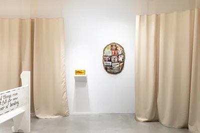 Installation view featuring a white church pew with black text, a gelatin sculpture containing a dried fish and an oval shaped textile sculpture hanging on the wall, surrounded by warm tan anti-microbial medical curtains.