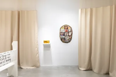 Installation view featuring a white church pew with black text, a gelatin sculpture containing a dried fish and an oval shaped textile sculpture hanging on the wall, surrounded by warm tan anti-microbial medical curtains.