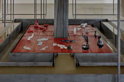 : Large sculpture with a red base that has smaller red, white, and black sculptures on the surface and large steel beams surrounding it forming a rectangular shape.