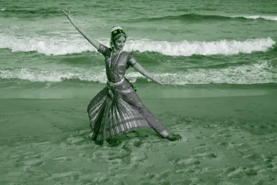 Performing artist Shriya Srinivasan poses in the sand on a beach with waves coming in behind her.