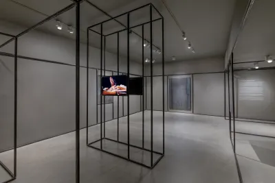 Gallery with grey walls and flooring featuring two monitors facing back-to-back on an open steel sculpture that runs throughout the gallery with two welded steel drawings in the background.
