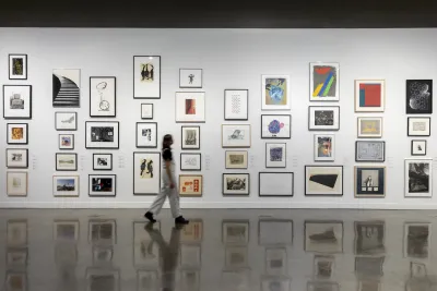 Installation view with framed artworks lining the wall and person in white overalls in motion walking through gallery.