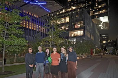 A group of six stand next to one another for a photo in a courtyard with two light sculpture above them in a dark courtyard.