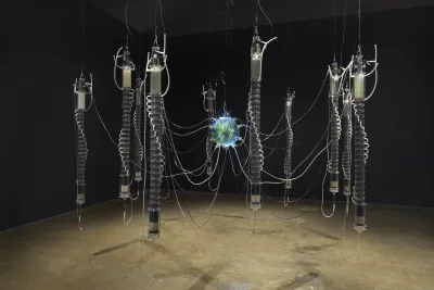 Dark gallery with a water processing system suspended from the ceiling with eleven chambers connected through various tubes and wires. At the center, a glass sphere contains livings plants, small fish, and processed wastewater.