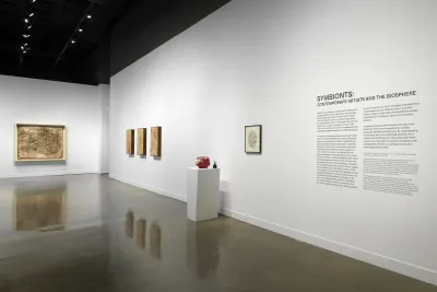 Black text on white title wall reads “SYMBIONTS: CONTEMPORARY ARTISTS AND THE BIOSPHERE” with show introduction. Work by Lin, Sutela, and Mobarak is in the background, reflected on the shiny concrete floor.