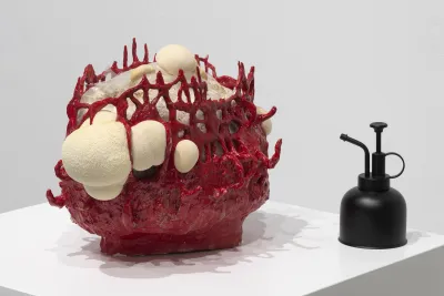 Sculpture of a lion’s mane mushroom that resembles a white foamy substance and grows inside a bright red ceramic vessel with a loose woven composition and many openings sits on a pedestal next to a matte black plant sprayer.