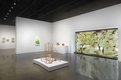 A beige lattice-like sculpture on a low white pedestal centers the gallery. A cluster of prints, a glowing green artwork, two small spherical sculptures, and a waxy window installation are in the background.