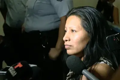 Video still of a woman with dark hair surrounded by a camera crew and microphones with her gaze to the left.