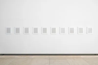 Ten grey plaster rectangles are framed in vertical, white wooden frames and hung equidistant in a horizontal line on a white wall. A thin, dark strip separates the wall and the beige tiled floor.