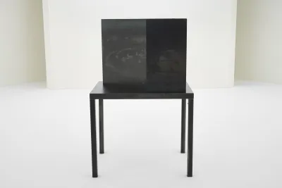 A square-looking black steel structure is on a four-legged table made of the same material in a gallery with white walls and a perfectly white floor.