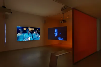 Two screens are situated at a right angle hanging in a room with an orange hue. On the left screen, three Asian women in white clothing look at one another. On the right screen a man gazes to the left.