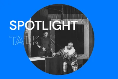 Bright blue background with white text that reads &quot;Spotlight Talk.&quot; In the center, there is a round blackened white image of a bald man wearing a fur collared coat sitting and a woman to his left standing behind a table. A microphone is suspended above them.