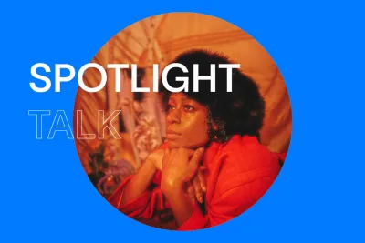 Bright blue background with white text that reads &quot;Spotlight Talk.&quot; In the center, there is a round image of a black woman wearing a fur hat and a red suite. She rests her hands on her chin and looks of into the distance to the left.