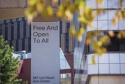 A large rectangular gray sign is visible through blurry leaves on a tree branch in the foreground. The text on the sign reads &quot;Free And Open To All MIT List Visual Arts Center&quot;