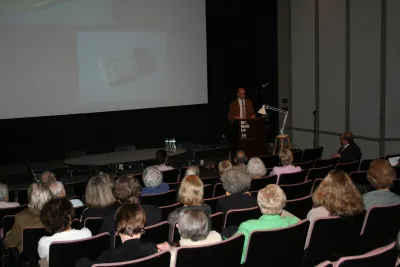 A speaker standing in front of a projector screen at a a black podium that has a sign that reads &quot;MIT Media Lab&quot;. There is a large crowd of people in the audience watching in the foreground of the image.