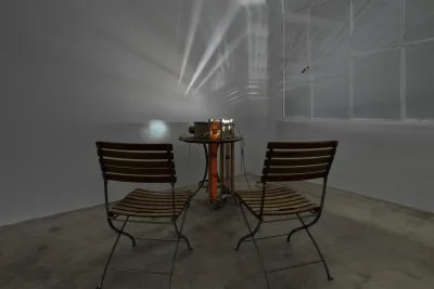 A pair of slatted wood folding chairs are clustered around a matching table that supports a homemade projector made of cardboard components and a bright orange band. Light spills from the top of the projector which also throws a glowing round orb containing scrolling text onto the wall.