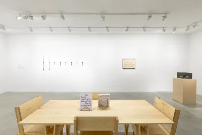 A white walled room with a tan wooden table and four tan wooden chairs in the middle of the room. To the right side of the table there is a wooden pedestal with a gray Plexiglas cube on top. There is a black framed drawing hung on the wall to the left of the pedestal. A row of eight test tubes are hung onto the wall to the left of the drawing.