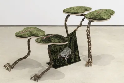 Four green crushed velvet panels each shaped like a trefoil spear to perch on top of a skeletal armature of sinewy braided bronze giving the overall impression of a four-legged animal crouching on its forelegs. At the animal’s belly, a flag of the same green cloth bears an embroidered design of a dog that appears to be either mid-gallop or hogtied.