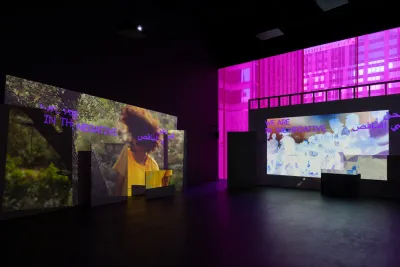 A dark room with two multi-channel video installations. On the right side of the image there is a large rectangular screen with two smaller rectangular panels positioned in front and pink light illuminates the background in a rectangular shape. On the left side of the image a large rectangular screen can be seen in front of a black background with two smaller rectangular panels positioned in front. The text within both video projections reads “WE ARE IN THE NEGATIVE”.