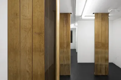 Series of three two-foot-wide floor-to-ceiling wood sculptures that appear to extend into the ceiling. Left side of image shows close up of scratched graffiti on the sculptures’ wooden planks.