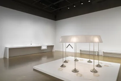 On a low pedestal in the foreground, seven standing lamps are conjoined by a single ring-shaped lampshade. A neat coil of pale cord sits near the base of each lamp. Behind it, small glass sculptures sit on a shelf, indistinct from the white wall behind them. A chevron-shaped wall piece is affixed to the wall nearby.