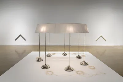 On a low pedestal in the foreground, seven standing lamps are conjoined by a single ring-shaped lampshade. A neat coil of pale cord sits near the base of each lamp. The sculpture is flanked, in the background, by two chevron-shaped wall pieces. The one on the left is dark, while the one on the right is pale.