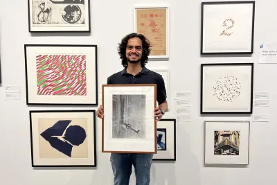 A student with brown hair holds a large framed artwork in front of a white gallery wall with numerous other framed works of art.