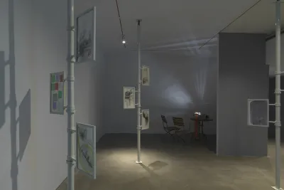 Three metal poles extend from floor to ceiling in a gallery. Two or three framed works on paper with layered compositions are attached to each one. The frames appear to be able to rotate around the poles. In the background on the left, a projector on a table casts an abstract light pattern across the wall, accompanied by two chairs.