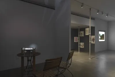 A makeshift projector sits on a wooden table, accompanied by two matching chairs. Two cords and a strip of material are attached to the device, which projects a faint circle of light onto the back wall. On the right and in the background, two floor-to-ceiling poles with drawings attached and a rectangular, more conventional projected image are visible.