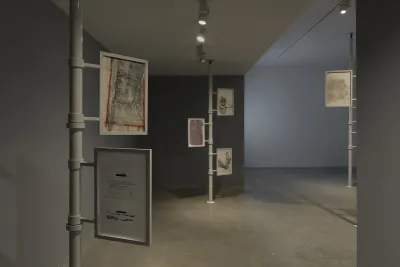Three metal poles extend from floor to ceiling. Two or three framed works on paper are attached to each one. Nearest to the camera, one work appears to be a printed document with some lines redacted; another is a drawing with a geometric layout resembling an architectural plan. The frames appear to be able to rotate around the poles.