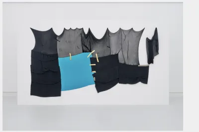 A large textile work hangs on a freestanding wall. It is composed of around nine sections of textile in varying shades of gray and black, seemingly “patched” together with strips of yellow fabric. One section is sky blue and stands out. The edges of the piece are irregular: sometimes, the fabric hangs freely in relatively straight lines; but along the top edge, the curved edge simply weight, tension, and stretching.